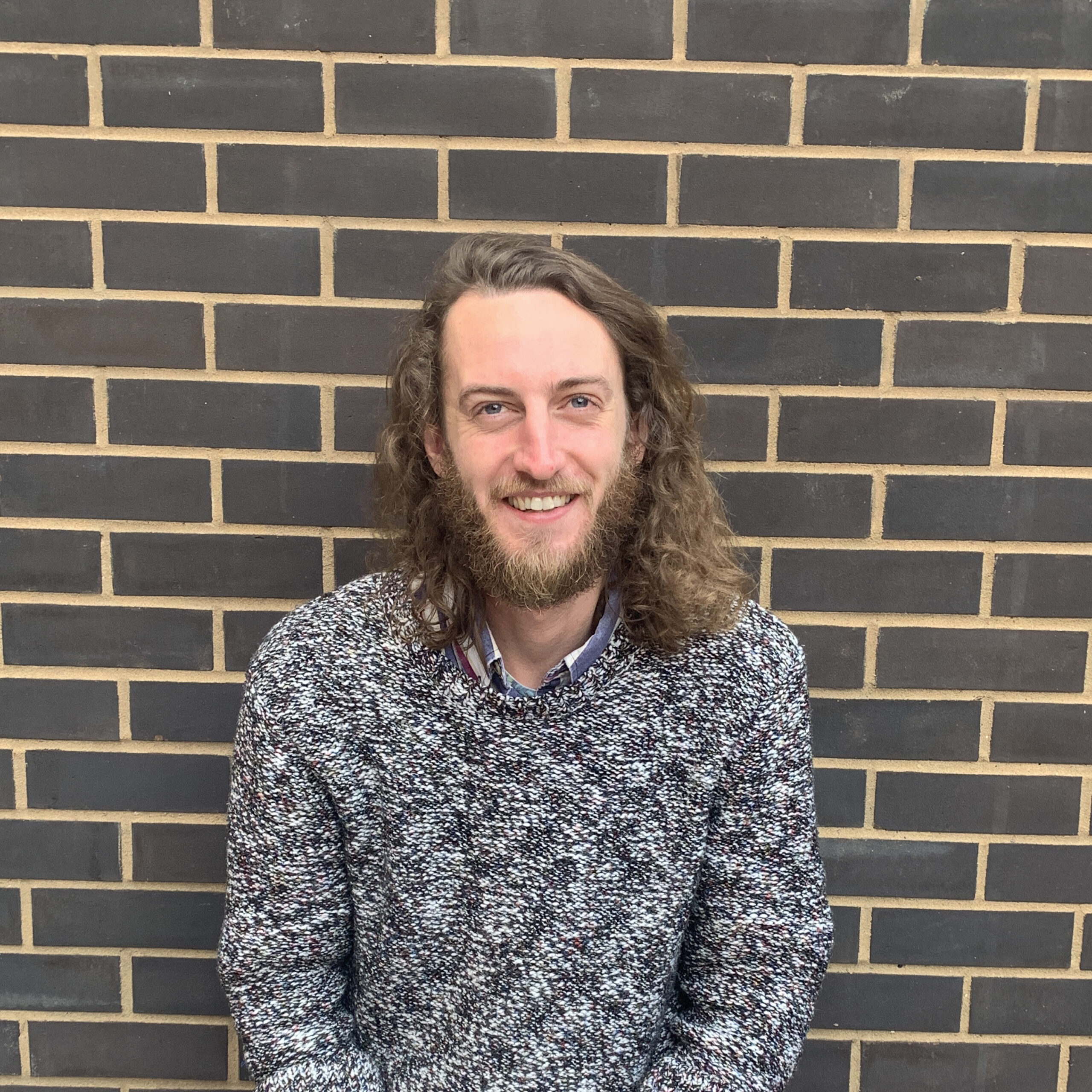 A medium shot of Rhys, an NN Ambassador. Rhys is white with shoulder length brown hair, wearing a grey and white speckled jumper and is smiling at the camera. Rhys is standing with a grey brick wall behind him.